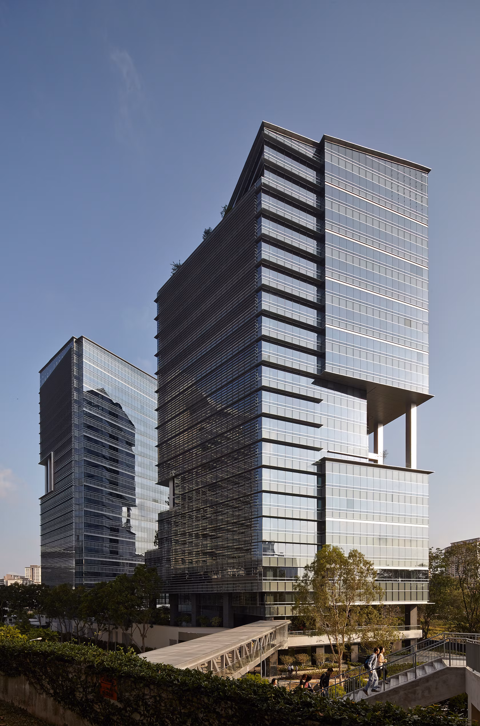 Two modern glass metropolis office buildings with reflective windows under a clear sky, with a footbridge and people walking nearby.